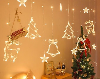 May include: A set of hanging LED string lights featuring illuminated shapes of stars, Christmas trees, bells, and reindeer. A sign with the text Merry Christmas is suspended on the wall, and a small decorated Christmas tree stands nearby.