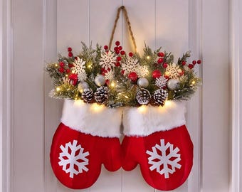 Puede incluir: Un par de manoplas de tela roja con aplicaciones de copos de nieve blancos, rematadas con un arreglo festivo de ramas de pino escarchadas, bayas rojas, adornos plateados, piñas y copos de nieve blancos, iluminadas por luces LED cálidas.