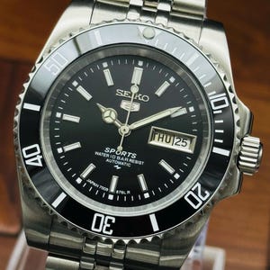 May include: A stainless steel Seiko 5 Sports automatic wristwatch with a black dial, a rotating black bezel with white numerals, and a silver-tone link bracelet. The dial displays the text Seiko 5 Sports, Water 10 Bar Resist, Automatic, and Japan 7009.