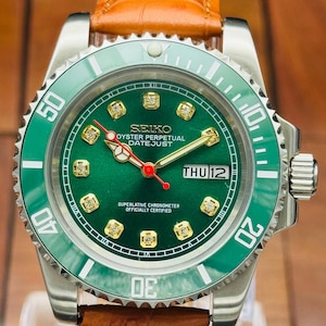 May include: A wristwatch with a green dial, gold-tone hour markers with embedded stones, and a green rotating bezel. The dial features the text SEIKO OYSTER PERPETUAL DATEJUST, a day-date window showing THU 12, and gold hands with a red second hand.