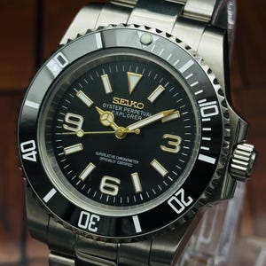 Pode incluir: Um relógio de pulso em aço inoxidável com mostrador preto, ponteiros e marcadores de hora dourados. O mostrador exibe o texto SEIKO, OYSTER PERPETUAL, EXPLORER e SUPERLATIVE CHRONOMETER OFFICIALLY CERTIFIED. Possui uma luneta giratória preta.