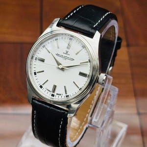 May include: A Jaeger-LeCoultre Club wristwatch with a round silver-tone case, a white dial featuring black hour markers, and silver hands. The watch includes a black leather strap with white stitching and the text T SWISS MADE T.
