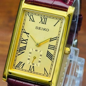 May include: A rectangular Seiko wristwatch with a gold-tone dial, black Roman numeral hour markers, and a sub-dial for seconds. The watch features a polished gold-tone case and a dark burgundy leather strap with a textured, embossed finish.