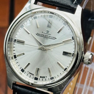 Può includere: Un orologio da polso Jaeger-LeCoultre Club color argento con quadrante circolare argento spazzolato, indici a bastone argentati e lancette argentate. L'orologio presenta un cinturino in pelle nera con cuciture bianche e una corona laterale.