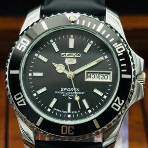 Pode incluir: Um relógio de pulso automático Seiko 5 Sports com mostrador preto, marcadores de hora luminosos e bisel rotativo preto. O relógio possui uma janela de dia e data na posição das 3 horas, ponteiros prateados e pulseira de couro preta. O mostrador exibe SEIKO, SPORTS, WATER 10 BAR RESIST, AUTOMATIC e JAPAN 7009 876L R.