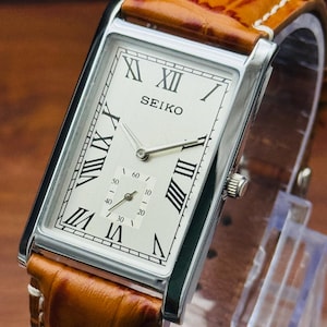 May include: A rectangular Seiko wristwatch featuring a silver-toned dial with black Roman numeral hour markers and a small seconds sub-dial at the bottom. The watch has a polished silver-colored metal case and a textured tan leather strap.