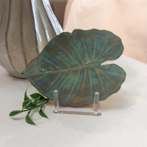 May include: A ceramic leaf-shaped dish featuring a teal and green glaze with visible brown veining and a textured surface. The dish is displayed upright on a clear acrylic stand, showcasing its organic form and detailed leaf structure.
