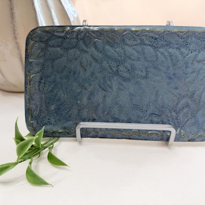 May include: A rectangular ceramic tray with a blue, textured glaze featuring a subtle embossed leaf pattern across the surface. The tray is displayed upright on a silver metal stand, showcasing its glossy finish and rounded corners.