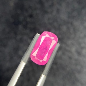 May include: A vibrant, hot pink gemstone with a rectangular cushion cut, featuring faceted surfaces that reflect light. The gemstone is held securely by a pair of metal tweezers, showcasing its saturated color and clear, geometric internal structure.