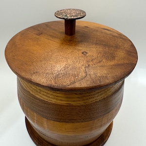 May include: A cylindrical wooden container with a lid featuring a circular copper-toned knob. The body is constructed from alternating horizontal bands of light and dark wood, showcasing a natural grain pattern and a smooth, polished finish.
