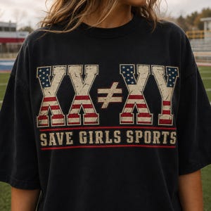 Könnte beinhalten: Ein schwarzes Kurzarm-T-Shirt mit einem abgenutzten Grafikdruck mit dem Text XX ≠ XY und SAVE GIRLS SPORTS. Die Buchstaben XX und XY sind mit einem rot-weiß-blauen amerikanischen Flaggenmuster gefüllt, der Text ist in fetter Blockschrift.