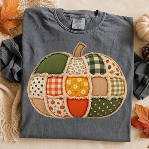 May include: A charcoal grey short-sleeve t-shirt featuring a large, central pumpkin graphic made of various fabric patches. The pumpkin design includes green, orange, and tan squares with patterns like gingham, polka dots, and floral prints.