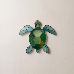 Può includere: Una decorazione da parete in vetro colorato a forma di tartaruga marina, con un guscio centrale composto da segmenti di vetro in varie tonalità di verde, circondato da quattro pinne in toni azzurro e verde acqua, delineate da linee di saldatura.