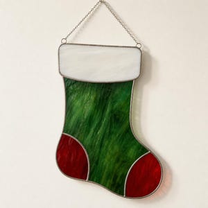 Puede incluir: Un adorno de calcetín navideño de vidriera con un puño blanco, un cuerpo verde texturizado y secciones rojas en el talón y la punta. La pieza está construida con líneas de soldadura de metal oscuro e incluye una cadena de metal para colgar.