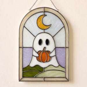 Puede incluir: Un atrapasoles de vidrieras en forma de arco que presenta un fantasma blanco sosteniendo una calabaza naranja. Sobre el fantasma hay una luna creciente amarilla. El fondo incluye paneles de vidrio morado claro y verde, enmarcados en metal oscuro.