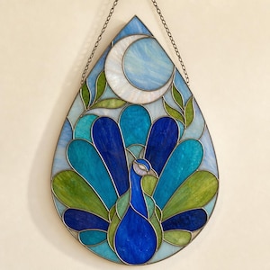 Op de afbeelding: Een druppelvormige glas-in-lood zonnevanger met een pauw met blauw en groen verenkleed. Boven de pauw bevindt zich een witte maansikkel tegen een lichtblauwe achtergrond. Het geheel is gevat in een metalen frame en hangt aan een ketting.
