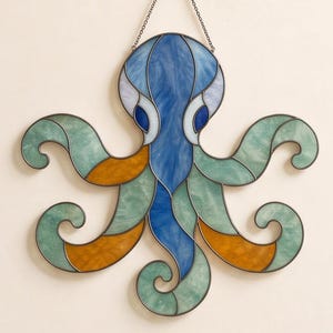 May include: A stained glass octopus suncatcher featuring a central blue body with dark blue eyes. The eight tentacles are crafted from segments of marbled seafoam green and amber glass, all held together by dark metal solder lines and a hanging chain.