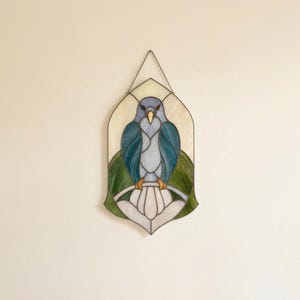 May include: A stained glass suncatcher featuring a blue bird perched on a white branch. The bird has light blue and teal wings, a yellow beak, and yellow feet. It is set against a cream background with green foliage elements and a metal chain.