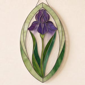 以下が含まれることがあります： 中央に紫色のアイリスの花と緑の葉が描かれた楕円形のステンドグラスパネル。デザインは淡い緑色と白色のガラスセグメントで縁取られ、暗い色の金属フレームで繋ぎ合わされています。上部には小さな金属製のチェーンが付いています。