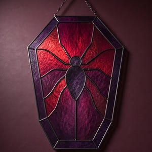 May include: A coffin-shaped stained glass panel featuring a central spider motif. The piece is constructed with dark purple and vibrant red textured glass segments held together by thin metal cames, suspended by a small metal chain at the top.