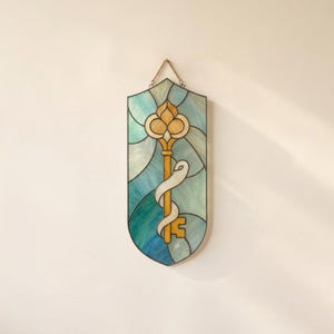 Op de afbeelding: Een schildvormig glas-in-loodpaneel met een centraal gouden sleutelontwerp omwikkeld met een wit lint. De achtergrond bestaat uit verschillende tinten blauw en groenblauw glas. Een metalen ketting is aan de bovenkant bevestigd om op te hangen.