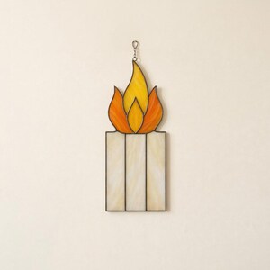 Peut inclure: Un attrape-soleil en vitrail en forme de bougie avec une flamme. La base de la bougie est composée de trois panneaux verticaux en verre texturé blanc cassé. La flamme au-dessus est faite de pièces de verre jaune et orange avec des soudures en métal.