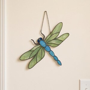 May include: A stained glass dragonfly suncatcher featuring four translucent light green wings with dark leaded outlines. The body is composed of several segments of bright blue glass, and the piece hangs from a thin metal chain.