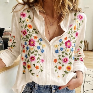 Könnte beinhalten: Ein weißes, langärmeliges Hemd mit Knopfleiste und farbenfroher Blumenstickerei. Das Design zeigt rosa, orange, gelbe und blaue Blüten mit grünen Stielen und Blättern, die über die Vorderseite und die Ärmel verteilt sind. Der Stoff ist strukturiert.