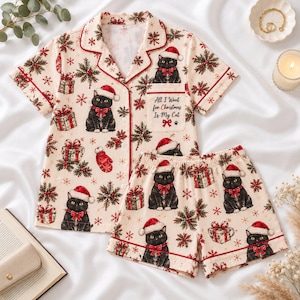 Può includere: Un set pigiama color crema a maniche corte e pantaloncini, caratterizzato da una stampa ripetuta di gatti neri con cappelli da Babbo Natale, regali rossi e fiocchi di neve verdi. La camicia ha una tasca con il testo All I Want for Christmas Is My Cat.