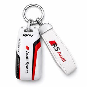 Peut inclure: Un étui de clé Audi Sport blanc avec des accents noirs et rouges, portant les inscriptions Audi, quattro et Audi Driving Experience. Il est attaché à une lanière en similicuir blanc avec le logo RS Audi rouge et noir sur un anneau en métal argenté.