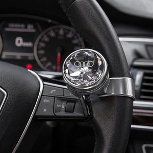 May include: A circular, faceted crystal steering wheel spinner knob featuring the silver Audi logo embedded inside. The accessory is mounted on a black leather-wrapped steering wheel with a metallic silver clip attachment.