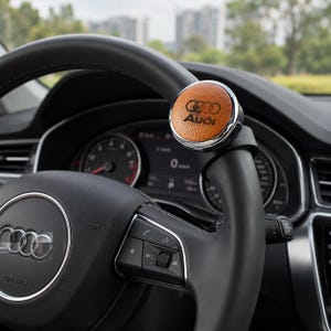 Op de afbeelding: Een ronde stuurwielknop van bruin leer met het zwarte Audi-logo en de tekst. Het accessoire is gemonteerd op een met zwart leer bekleed stuurwiel met een zilveren sierring, geplaatst in het interieur van een voertuig.