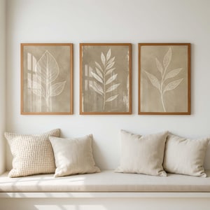 May include: Three framed wall art prints featuring white line drawings of botanical leaves on a beige background. Each print is encased in a light brown wooden frame. The artwork is displayed in a horizontal row above a bench with four beige pillows.