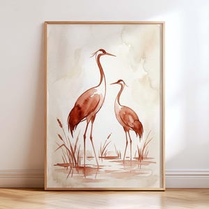 May include: A framed watercolor print featuring two rust-colored cranes standing in shallow water among reeds. The artwork uses a soft, neutral background with varying shades of brown and beige, depicting the birds in a serene, natural setting.