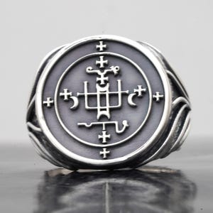May include: A silver ring featuring a circular face with a dark grey recessed background. The centre displays an intricate, raised silver sigil design composed of crosses, crescent shapes, and geometric lines. The band has a smooth, polished finish.