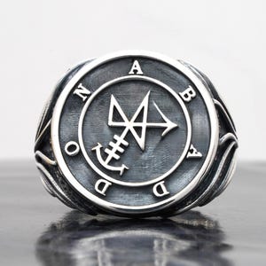 May include: A silver ring featuring a circular face with an oxidised finish. The centre displays a complex geometric sigil, surrounded by the letters A, B, A, D, O, N arranged in a circle. The band has decorative side detailing with a polished edge.