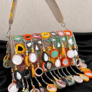 Puede incluir: Un bolso rectangular con una superficie vibrante y multicolor adornada con numerosos espejos pequeños, abalorios intrincados y dijes de espejo colgantes envueltos en tela en tonos amarillo, naranja, rosa, verde y gris con correa.