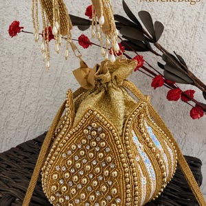 May include: A gold drawstring pouch bag featuring intricate beadwork, metallic sequins, and clear crystals. The bag has a structured petal-like design, a gold braided handle, and a dangling beaded fringe. The text NuvelleBags is visible.