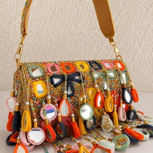 Puede incluir: Un bolso rectangular con intrincados abalorios dorados, espejos cuadrados y dijes de espejo colgantes envueltos en tela de colores naranja, amarillo, rosa, azul y verde. El bolso incluye una correa de hombro a juego y herrajes de metal dorado.
