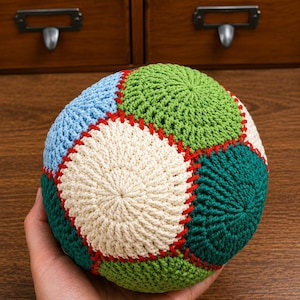May include: A spherical crochet ball constructed from multiple circular panels in shades of light blue, cream, light green, and dark green. The panels are joined together with visible red yarn stitching, creating a textured, multi-colored surface.