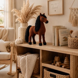 May include: A brown crocheted horse toy with a black yarn mane and tail, standing on four legs with black hooves. The horse has a textured, stitched surface and is positioned on a wooden surface alongside books and decorative items.
