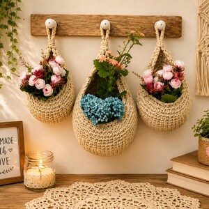 May include: Three beige crochet hanging baskets suspended from a wooden rack, each containing artificial flowers. The baskets are teardrop-shaped with a thick, textured weave. A framed sign reads "HANDMADE with LOVE" next to a candle in a glass jar.