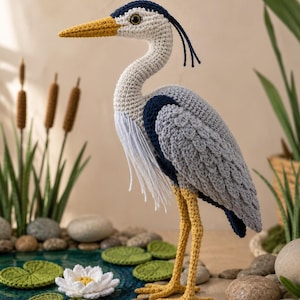 May include: A crocheted heron figurine featuring a grey, white, and dark blue body with a yellow beak and long yellow legs. The bird has a white fringed chest, a dark blue crest, and a single brown eye. It stands on a surface with crochet lily pads.