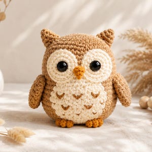 May include: A crocheted owl figurine featuring a light brown body, a cream-coloured chest with small brown V-shaped patterns, large black plastic eyes, a small orange beak, and two small orange feet. The owl has pointed ear tufts and rounded wings.