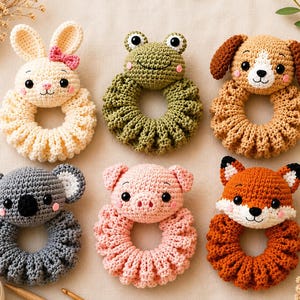 May include: A set of six crocheted animal rattles featuring a cream rabbit with a pink bow, a green frog, a tan dog, a gray koala, a light pink pig, and an orange fox. Each animal head is attached to a circular, textured, crocheted ring base.