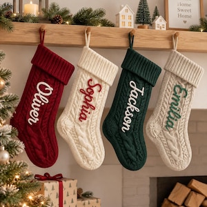 May include: Four cable-knit Christmas stockings hanging in a row. The first is red with white text reading Oliver, the second is cream with red text reading Sophia, the third is dark green with white text reading Jackson, and the fourth is cream with green text reading Emilia.