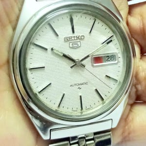 May include: A silver-tone Seiko 5 automatic wristwatch with a textured white dial, silver baton hour markers, and a date window displaying the number 28 in red and black. The watch features a stainless steel link bracelet and a round case.