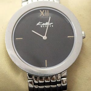 May include: A silver-tone wristwatch with a round case and a dark gray dial. The watch face features the text Kolber Geneve, a Roman numeral XII at the top, and simple stick markers. It includes a multi-link metal bracelet with a polished finish.
