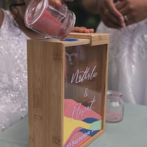 Peut inclure: Un cadre de cérémonie de sable rectangulaire en bambou avec une façade transparente portant le texte blanc Nathalie & Florent et 20 Septembre 2025. Le cadre est partiellement rempli de couches de sable rose, jaune et bleu, avec un pot en verre.