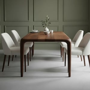 May include: A rectangular wooden dining table with rounded corners and four legs, accompanied by four upholstered chairs with white fabric seats and dark wooden legs. A small white vase with green leaves sits on the center of the table.